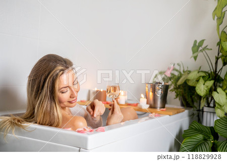 A young beautiful woman lies in a bath with foam and rose petals. Drinks champagne and white wine.  118829928