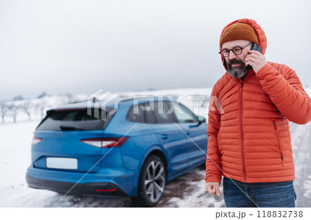Angry man standing by electric car, battery run out of power before reaching destination. Man phone calling for help waiting for breakdown service car, tow truck. 118832738