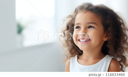 Portrait of a charming young girl with dark curly hair smiling against a bright background 118833460