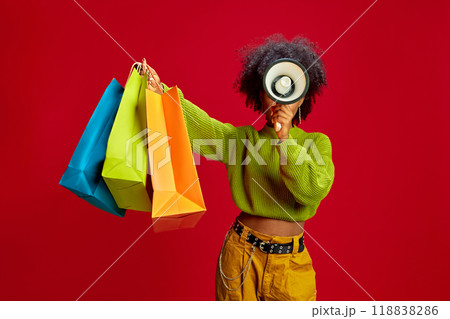 Lifting colorful bags and using megaphone, woman promotes consumerism in fun, engaging way against bold red background. Lifting colorful bags and using megaphone, woman promotes consumerism in fun, engaging way against bold red background. 118838286