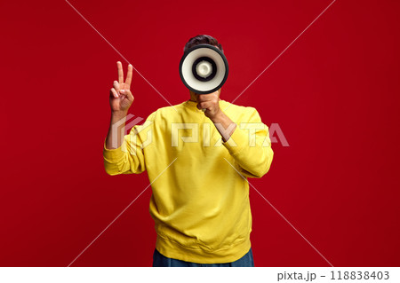 Person in bright yellow sweatshirt holds megaphone to face, making peace sign with their left hand against bright red background. Person in bright yellow sweatshirt holds megaphone to face, making peace sign with their left hand against bright red background. 118838403