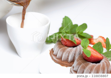 Chocolate fudge garnished with half a strawberry with petals. Garnished with sprigs of mint and strawberries. Nearby, they pour coffee into a cup. On the white surface of the table. 118838807