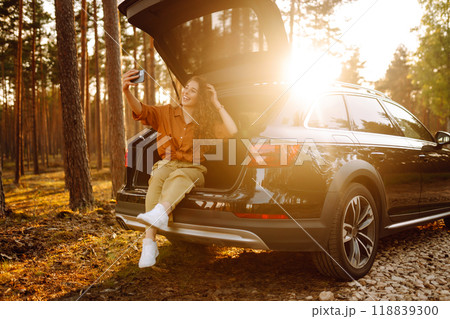 Happy stylish female tourist in trunk of a car with phone, taking a selfie, talking on a video call. 118839300
