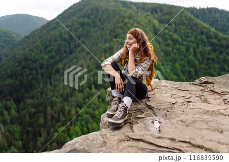 Beautiful traveler with a yellow hiking backpack observes the mountain scenery from a cliff. 118839590