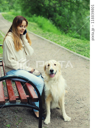 Young woman owner talking on phone with Golden Retriever dog sitting on bench in summer park Young woman owner talking on phone with Golden Retriever dog sitting on bench in summer park 118839801
