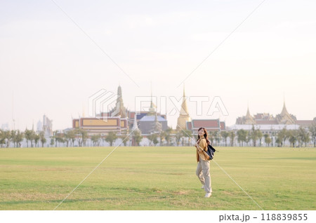 Traveler asian woman in her 30s, backpack slung over her shoulder, explores Wat Pra Kaew with childlike wonder. Sunlight dances on the golden rooftops, and a carefree smile 118839855