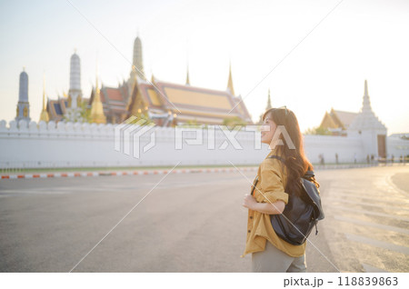 Traveler asian woman in her 30s, backpack slung over her shoulder, explores the intricate details of Wat Pra Kaew with childlike wonder. Sunlight dances on the golden rooftops. 118839863