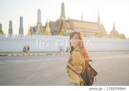 Traveler asian woman in her 30s, backpack slung over her shoulder, explores the intricate details of Wat Pra Kaew with childlike wonder. Sunlight dances on the golden rooftops. Traveler asian woman in her 30s, backpack slung over her shoulder, explores the intricate details of Wat Pra Kaew with childlike wonder. Sunlight dances on the golden rooftops. 118839864