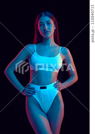 Confident woman in white sports bra and high-waisted briefs poses in vibrant neon light against black studio background. Confident woman in white sports bra and high-waisted briefs poses in vibrant neon light against black studio background. 118840715