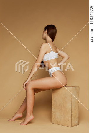 Female model poses seated on block, dressed underwear creating natural flow of lines against beige studio background. 118840726