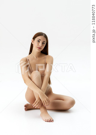 Seated with one leg bent, woman gazes softly at camera against white studio background. Her serene expression and relaxed pose create calm atmosphere 118840773