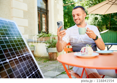 Young man charging smartphone from solar panel while sitting in cafe. Handsome male drinking coffee and checking his phone next to solar battery. Conscious male choosing sustainable lifestyle. 118841554