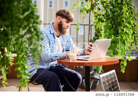 Man works on laptop and smartphone at outdoor table, with glass of iced coffee. Solar panel nearby, emphasizing sustainable, modern eco-friendly workspace that combines technology and sustainability. Man works on laptop and smartphone at outdoor table, with glass of iced coffee. Solar panel nearby, emphasizing sustainable, modern eco-friendly workspace that combines technology and sustainability. 118841566
