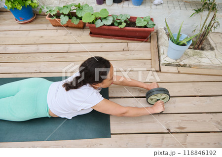 Woman exercising with ab roller on a wooden deck surrounded by plants and potted flowers, practicing fitness and core strengthening outdoors. Woman exercising with ab roller on a wooden deck surrounded by plants and potted flowers, practicing fitness and core strengthening outdoors. 118846192