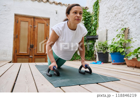 Determined woman performing push-ups with push-up bars on yoga mat in serene outdoor setting 118846219