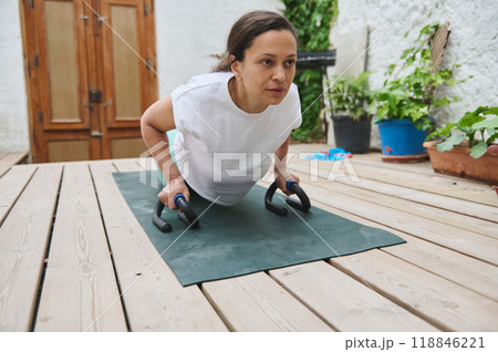 Woman practicing push-ups at home using push-up bars on a wooden deck for a healthy outdoor fitness routine 118846221