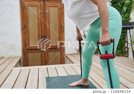 Woman exercising outdoors with resistance bands performing a bent-over row on a wooden deck in casual athletic wear. Woman exercising outdoors with resistance bands performing a bent-over row on a wooden deck in casual athletic wear. 118846234
