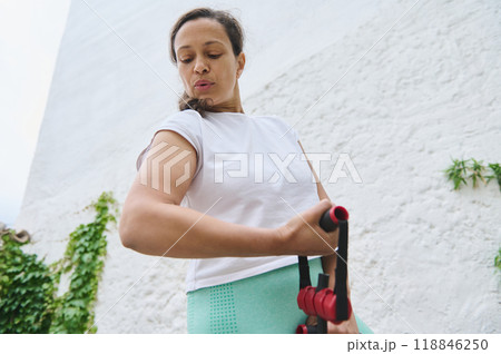 Woman exercising outdoors with resistance bands in a bright white and green environment, focused on her fitness routine 118846250