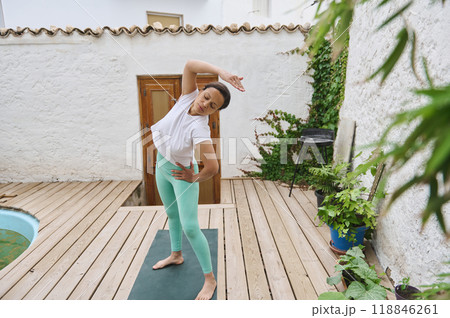 Woman practicing yoga outdoors on wooden deck surrounded by greenery, performing side stretch in peaceful setting 118846261