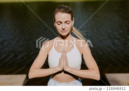 High angle shot of blonde Caucasian woman meditating in Pranamasana pose in park near river High angle shot of blonde Caucasian woman meditating in Pranamasana pose in park near river 118846912