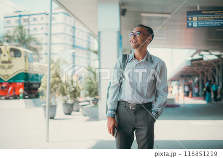 A confident man smiles at a bustling train station, embodying adventure and exploration 118851219
