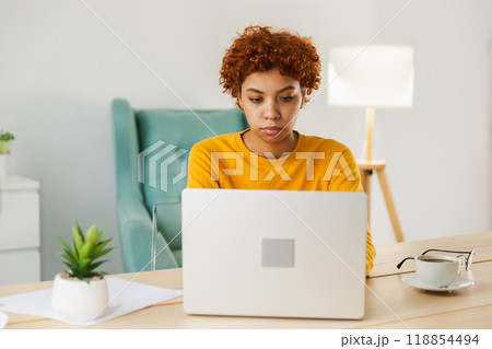 African american girl using laptop at home office looking at screen typing chatting reading writing email. Young woman having virtual meeting online chat video call conference. Work learning from home African american girl using laptop at home office looking at screen typing chatting reading writing email. Young woman having virtual meeting online chat video call conference. Work learning from home 118854494