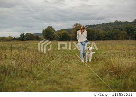 A Woman Taking a Leisurely Walk with Her Dog in a Scenic Field Beneath a Cloudy Sky A Woman Taking a Leisurely Walk with Her Dog in a Scenic Field Beneath a Cloudy Sky 118855512