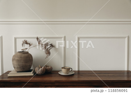 Elegant sideboard autumn arrangement. Stylish wooden sideboard dresser with minimalist accents. Clay vase, dry fern leaf, pumpkins, cup of coffee. Modern interior design trends, white wall background. 118856091