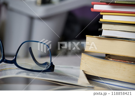 Reading glasses placed on an open book next to a stack of books, symbolizing a study or educational Reading glasses placed on an open book next to a stack of books, symbolizing a study or educational 118858421
