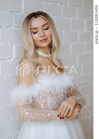 Close-up portrait blonde in a white fluffy wedding dress poses against a white brick wall. 118861486