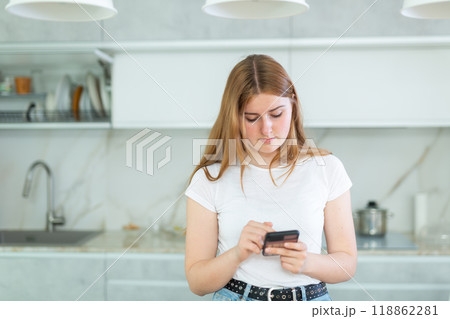 bored young woman stands in kitchen leans on countertop and surfs internet.Home pastime 118862281