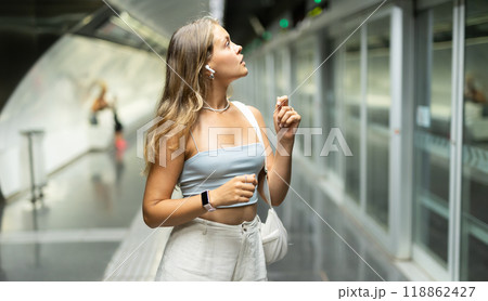 Girl with earphones is standing on subway platform, waiting for train to arrive 118862427