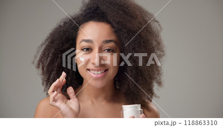 Beauty and healthcare concept - beautiful African American woman with curly afro hairstyle and clean, healthy skin applying moisturizer to her face with a slight smile and positive mood 118863310