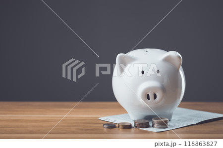 White piggy bank, bank book, and a pile of coins rest on a wooden table against a dark gray background. The scene symbolizes financial planning, savings, and the importance of managing money wisely. White piggy bank, bank book, and a pile of coins rest on a wooden table against a dark gray background. The scene symbolizes financial planning, savings, and the importance of managing money wisely. 118863827