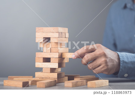 Businessman contemplates his next move in a stacking tower block game on a table against a gray wall background. Symbolizes strategic thinking, planning, and decision-making in business and life Businessman contemplates his next move in a stacking tower block game on a table against a gray wall background. Symbolizes strategic thinking, planning, and decision-making in business and life 118863973