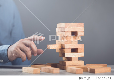 Businessman contemplates his next move in a stacking tower block game on a table against a gray wall background. Symbolizes strategic thinking, planning, and decision-making in business and life Businessman contemplates his next move in a stacking tower block game on a table against a gray wall background. Symbolizes strategic thinking, planning, and decision-making in business and life 118863974