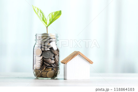 Glass jar filled with coins and a small seedling in the center, with stacked coins arranged from smallest to largest and a wooden house model on a table. Saving for house and financial growth Glass jar filled with coins and a small seedling in the center, with stacked coins arranged from smallest to largest and a wooden house model on a table. Saving for house and financial growth 118864103