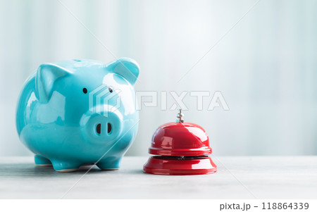 A close-up of a red service bell next to a piggy bank on a wooden table against a bright white background. The image represents a call to action for financial planning and savings. 118864339