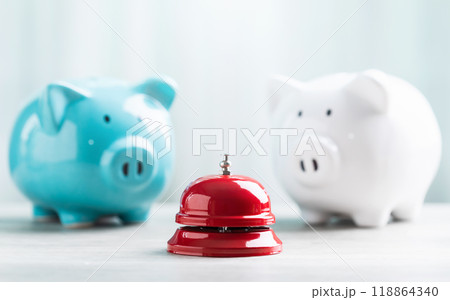 A close-up of a red service bell next to a piggy bank on a wooden table against a bright white background. The image represents a call to action for financial planning and savings. 118864340