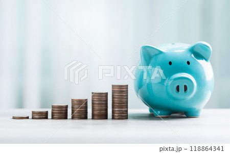 A blue piggy bank and stack of coins are neatly arranged on a table against a bright white background. The image represents the concept of saving money for the future. A blue piggy bank and stack of coins are neatly arranged on a table against a bright white background. The image represents the concept of saving money for the future. 118864341