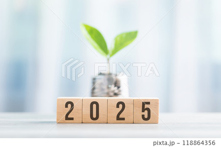 2025 is written on a wooden block next to a glass jar filled with coins, placed on a wooden table against a bright white. symbolizes financial planning, savings, and effective money management 118864356