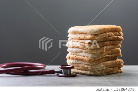 Slices of bread stacked together with a red stethoscope on a gray wooden table against a dark gray background. The image conveys the concept of the relationship between diet and health. Slices of bread stacked together with a red stethoscope on a gray wooden table against a dark gray background. The image conveys the concept of the relationship between diet and health. 118864379
