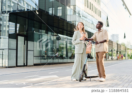 Wide shot of Caucasian woman holding cup of coffee and her Black male colleague with e-scooter chatting while standing outdoors Wide shot of Caucasian woman holding cup of coffee and her Black male colleague with e-scooter chatting while standing outdoors 118864581