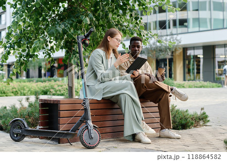 Wide shot of two young ethnically diverse coworkers sitting outdoors looking at digital tablet screen and discussing project during coffee break 118864582