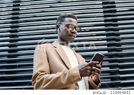 Low angle view of confident young Black man in formalwear listening to music via earbuds and texting message on smartphone while standing outdoors Low angle view of confident young Black man in formalwear listening to music via earbuds and texting message on smartphone while standing outdoors 118864681