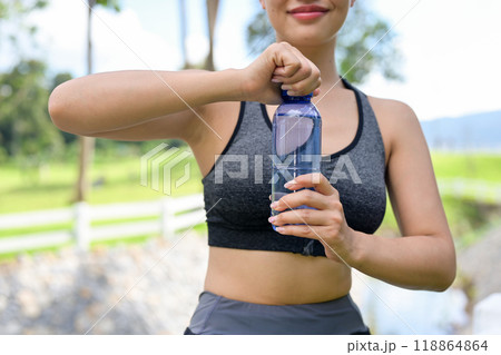 A fit Asian woman in sportswear opening a bottle of water and drinking after jogging in a park. 118864864