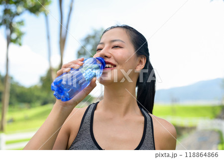 A fit woman in sportswear drinks from a water bottle, quenching her thirst after jogging in a park. A fit woman in sportswear drinks from a water bottle, quenching her thirst after jogging in a park. 118864866