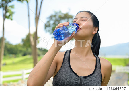 A fit woman in sportswear drinks from a water bottle, quenching her thirst after jogging in a park. A fit woman in sportswear drinks from a water bottle, quenching her thirst after jogging in a park. 118864867