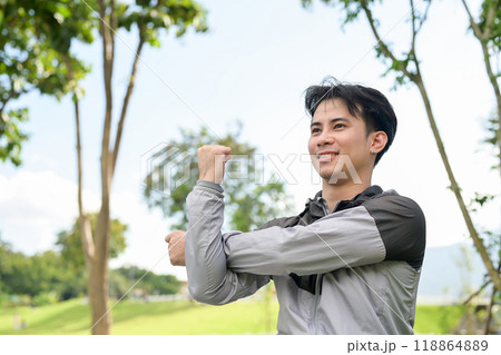 A handsome, fit Asian man in sportswear stretches his arm, warming up before jogging in a park. 118864889