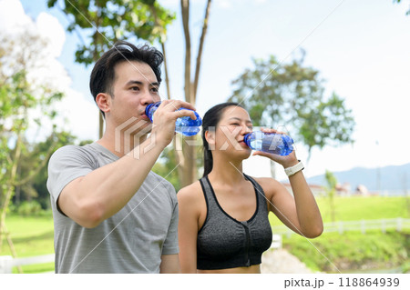 A thirsty Asian couple drinks water from their bottles while resting together after jogging. A thirsty Asian couple drinks water from their bottles while resting together after jogging. 118864939